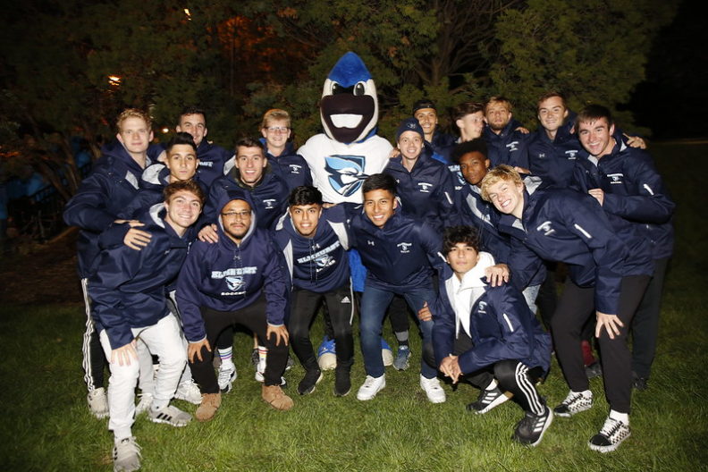 An athletic team poses with Elmhurst University mascot Victor E. Bluejay for a photo during a nighttime Homecoming pep rally.