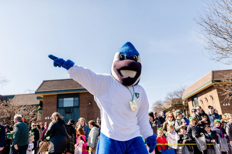 Elmhurst University mascot Victor E. Bluejay points to his right.