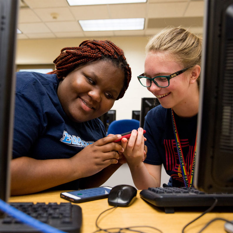 A Black female student and a white female student at Elmhurst University smile at each other between their computer terminals.