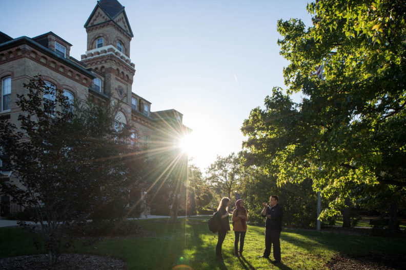 Students stand on the Elmhurst University campus with the Old Main building in the background and brilliant sunshine behind them.