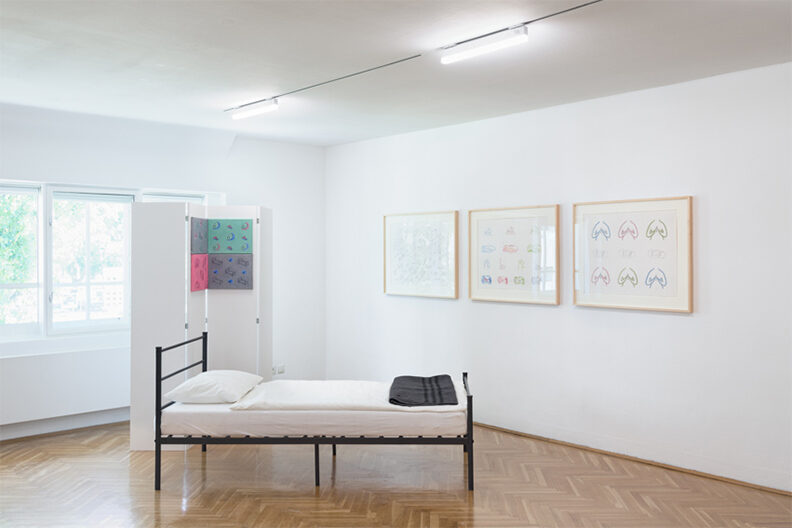 An art gallery room with an empty bed in the foreground and works of art hanging on the wall in the background.