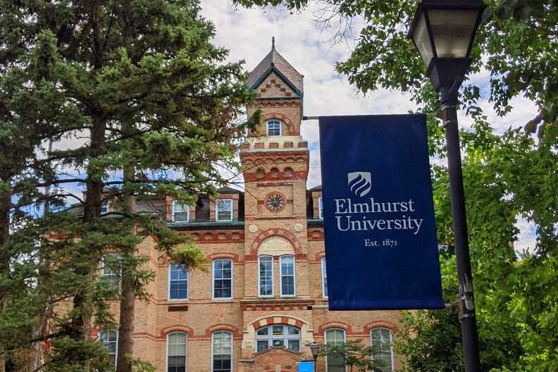 A banner reading "Elmhurst University" stands in the foreground, with the campus' Old Main building rising behind it.