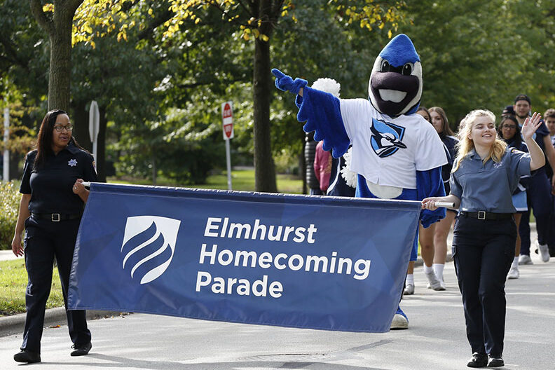 Two women hold a blue banner that reads "Elmhurst Homecoming Parade" while Elmhurst University mascot Victor E. Bluejay marches behind it.