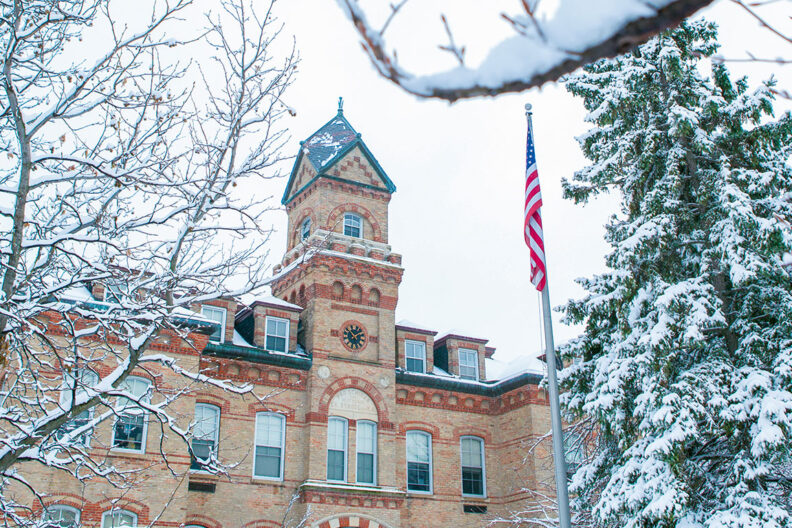 A photo of snow-covered tree branches in the foreground, with the front and bell tower of the Old Main building in the background on the campus of Elmhurst University.