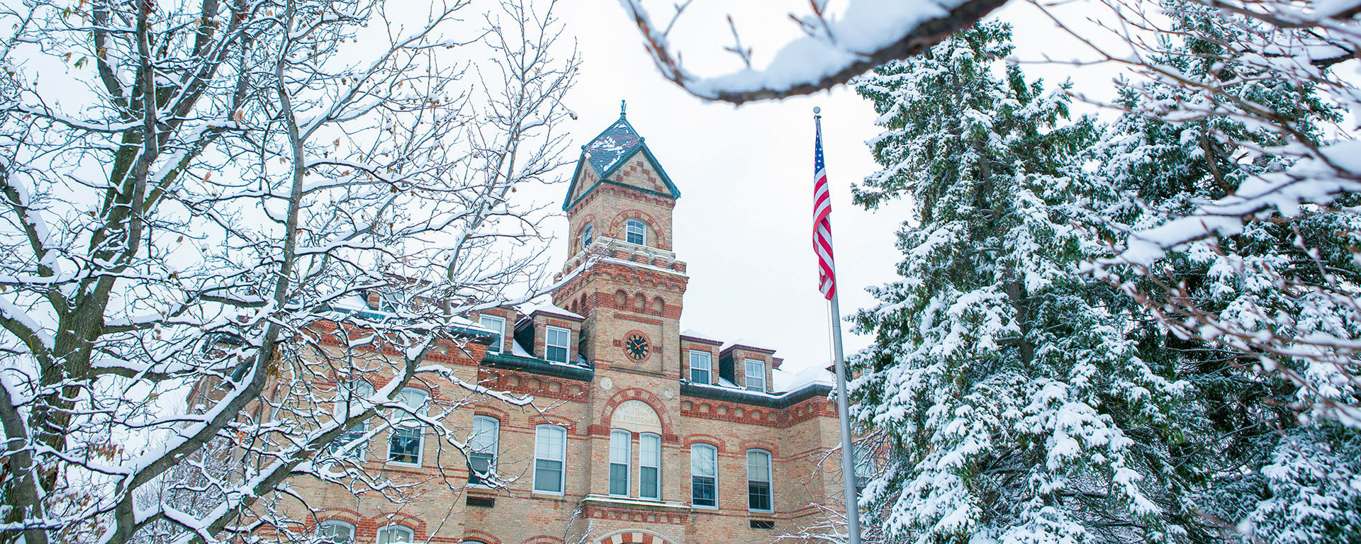 A photo of snow-covered tree branches in the foreground, with the front and bell tower of the Old Main building in the background on the campus of Elmhurst University.