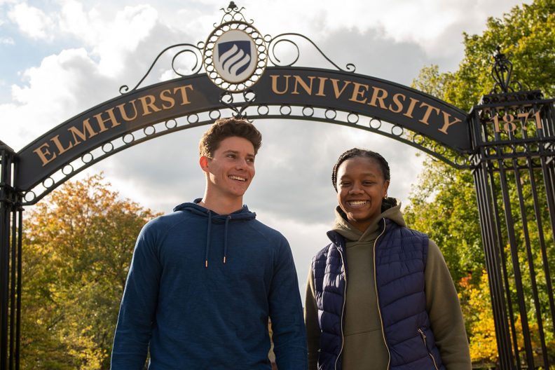 Two Elmhurst University students, one male, one female, smile underneath the campus Gates of Knowledge.