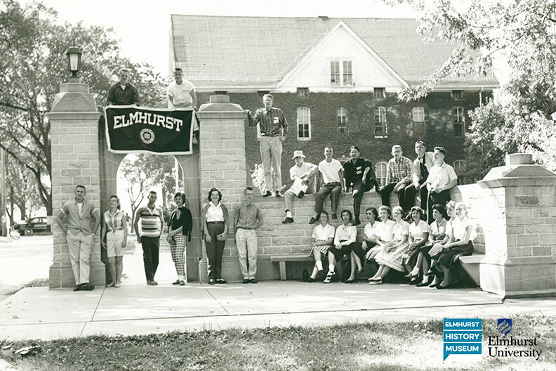 A black-and-white photo of students posing for a group photo at the entrance gates of Elmhurst University.