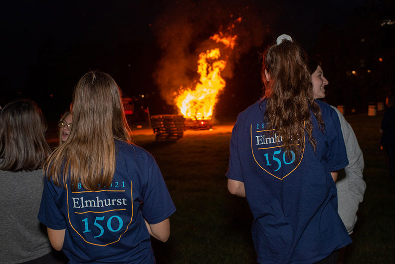 Two female students wearing "Elmhurst 150" T-shirts watch a roaring bonfire during Elmhurst University's 2021 Homecoming pep rally.