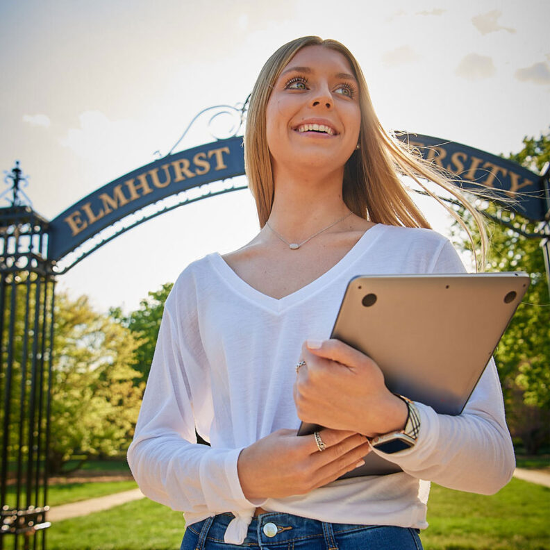 A female Elmhurst University student holding a laptop stands in front of the "Gates of Knowledge" on campus.