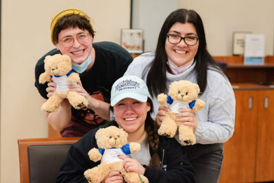 A male and two female Elmhurst University students smile while holding up Elmhurst 150th anniversary Build a Bear stuffed animals.