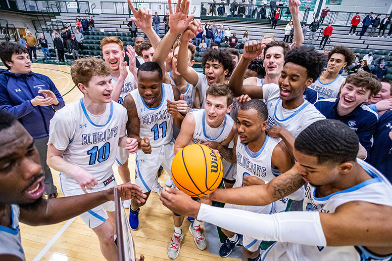 The Elmhurst University men's basketball team celebrates its CCIW conference championship victory in a huddle around a basketball in February of 2022.