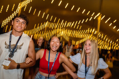 A male and two female Elmhurst University students link arms under a canopy of yellow lights during New Student Orientation in 2022.