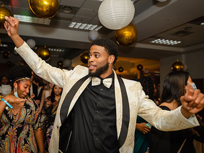 A Black male Elmhurst University student in a white tuxedo jacket, white bowtie and black dress shirt dances during the Presidents' Ball event.