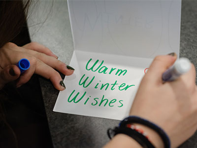 A student writes the message "Warm Winter Wishes" in green marker on a white card.