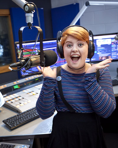 A female radio DJ sits at the microphone inside Elmhurst University's WRSE radio station broadcast booth.