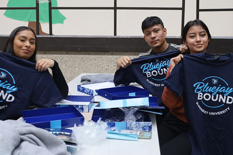 A male and two female students at Benito Juarez Community Academy holding up navy blue T-shirts that show they were accepted into Elmhurst University.