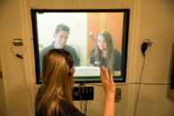 An Elmhurst University Communication Sciences and Disorders student waves from inside a sound booth at two other students on the other side of the glass.