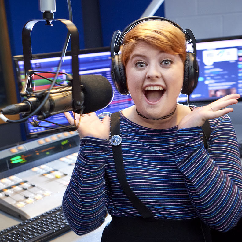 A female radio DJ sits at the microphone inside Elmhurst University's WRSE radio station broadcast booth.