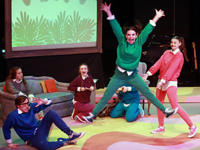 A scene from the Elmhurst University Theatre production of "Schoolhouse Rock Live," with a student-actress jumping up with arms and legs outstretched.