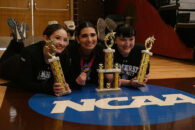 three girls holding three gold trophies by the NCAA seal